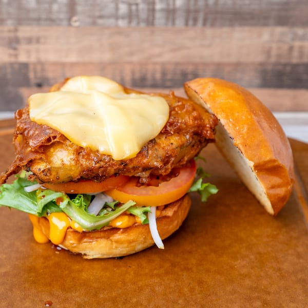 A fried chicken burger with melted cheese, lettuce, tomato, and sauce on a wooden board.