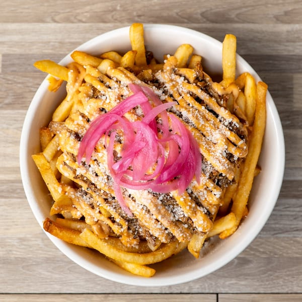 A white bowl of The Standing Room's loaded fries, topped with cheese, meat, and pickled red onions.