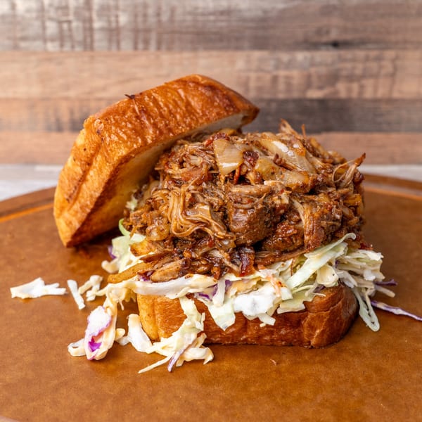 A pulled pork sandwich with coleslaw on a toasted bun, against a rustic wooden background.